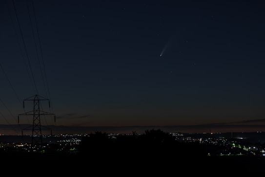 A night sky featuring a visible comet with a bright tail, above a cityscape with twinkling lights on the horizon. A silhouetted power line tower is visible on the left side, indicating a rural or suburban area.