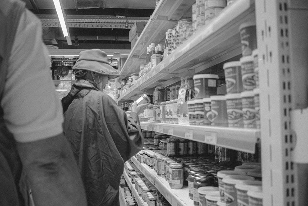 grayscale photo of man in jacket standing in front of shelf