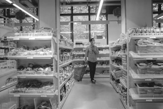 A person wearing a mask is walking down an aisle in a grocery store, pushing a shopping basket. Shelves on both sides are stocked with various packaged goods, and a large window at the back lets in natural light.