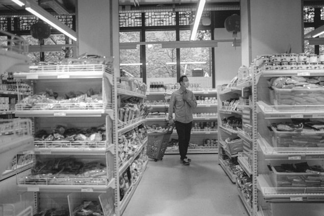 A person wearing a mask is walking down an aisle in a grocery store, pushing a shopping basket. Shelves on both sides are stocked with various packaged goods, and a large window at the back lets in natural light.