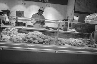 A butcher is working behind a glass counter filled with various cuts of meat. The butcher appears to be checking or preparing something in his hands. The background consists of white tiled walls with utensils hanging and a chart on display. The scene is monochromatic and has a somewhat vintage feel.