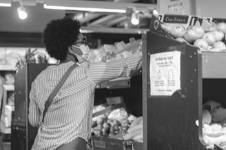 A person wearing a face mask and glasses is reaching for produce in a grocery store. The individual is dressed in a striped shirt with a shoulder bag draped across their back. Various fruits and vegetables are displayed in bins, and a sign with instructions is visible on the shelving unit.