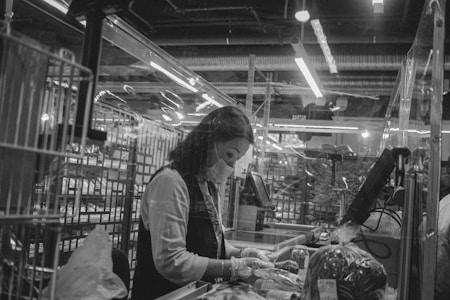 A person wearing a protective mask and gloves is working at a checkout counter in a store. The setting includes various items on the conveyor belt and a cash register. The environment appears industrial, with visible lighting fixtures and ventilation pipes above.
