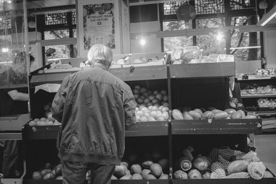 A black and white image of a person standing in front of a fruit display in a grocery store. The individual is wearing a jacket and appears to be selecting produce from the shelves, which are stocked with various fruits like apples and melons. There is signage in the background, along with other grocery items visible on adjacent shelves.