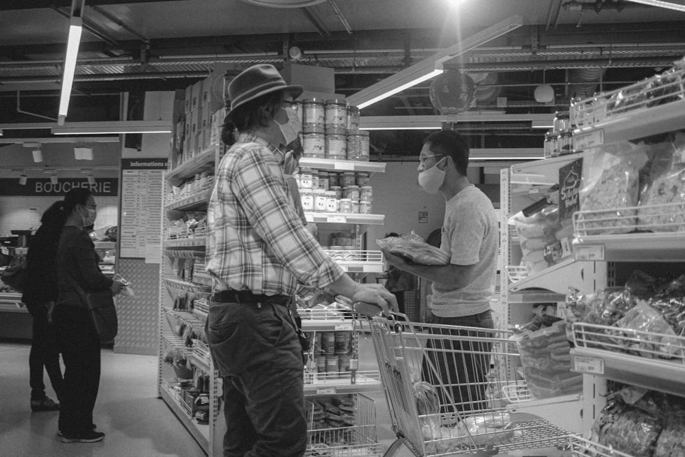 grayscale photo of man in plaid shirt and hat holding shopping cart
