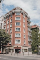 A corner building with a classic European architectural style, featuring red brick exterior walls and multiple white-framed windows. The lower level hosts a store named 'Asia Center' with signs in various languages. The building is surrounded by urban streets with trees and a section of the wall is covered in green ivy.