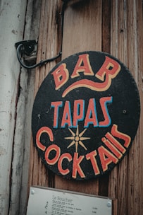 A vintage-style sign with bold, colorful lettering advertising a bar, tapas, and cocktails. The sign is circular and attached to a weathered wooden surface, alongside a mounted menu listing various dishes and prices.