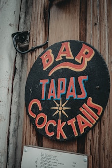 A vintage-style sign with bold, colorful lettering advertising a bar, tapas, and cocktails. The sign is circular and attached to a weathered wooden surface, alongside a mounted menu listing various dishes and prices.