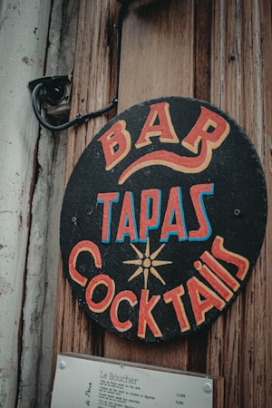 A vintage-style sign with bold, colorful lettering advertising a bar, tapas, and cocktails. The sign is circular and attached to a weathered wooden surface, alongside a mounted menu listing various dishes and prices.