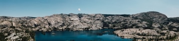 A panoramic view of a serene lake surrounded by rugged mountains and dense forest. The clear blue sky enhances the tranquil atmosphere. The water is calm, reflecting the surrounding natural beauty.