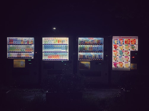 A row of illuminated vending machines displays a variety of drinks and snacks. The area around the machines is dark, emphasizing the bright electric lights and colorful product images. The setting appears to be outdoors, possibly in a quiet or dimly lit location.