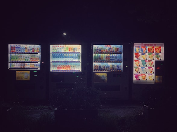A row of illuminated vending machines displays a variety of drinks and snacks. The area around the machines is dark, emphasizing the bright electric lights and colorful product images. The setting appears to be outdoors, possibly in a quiet or dimly lit location.