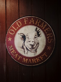 A circular vintage sign with a smiling pig illustration in the center. The sign reads 'Old Farmers Meat Market' in bold letters. The background is a dark wooden texture, adding a rustic feel to the overall look.