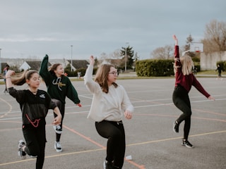 A group of four young people are engaging in an outdoor activity on a sports court. They are lifting their arms and appear to be mid-motion, suggesting they are dancing or exercising together. The background includes greenery, trees, and a basketball hoop, indicative of a recreational setting.