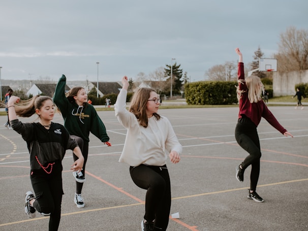 A group of four young people are engaging in an outdoor activity on a sports court. They are lifting their arms and appear to be mid-motion, suggesting they are dancing or exercising together. The background includes greenery, trees, and a basketball hoop, indicative of a recreational setting.