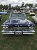 A classic vintage car is parked on a grassy field, surrounded by other antique cars and people. The car has a shiny navy blue exterior with chrome details, and a maroon license plate indicating it might be from Victoria. The car show is taking place under an overcast sky, with trees and advertisement signs visible in the background.