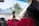 A person inside a car is taking a photo or video with a smartphone towards the outside. The individual is wearing a graduation cap and a red outfit with floral patterns. Outside, palm trees are visible, suggesting a sunny location. A banner with the word 'GRADUATES' is visible through the windshield.