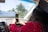 A person inside a car is taking a photo or video with a smartphone towards the outside. The individual is wearing a graduation cap and a red outfit with floral patterns. Outside, palm trees are visible, suggesting a sunny location. A banner with the word 'GRADUATES' is visible through the windshield.