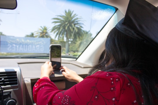 A person inside a car is taking a photo or video with a smartphone towards the outside. The individual is wearing a graduation cap and a red outfit with floral patterns. Outside, palm trees are visible, suggesting a sunny location. A banner with the word 'GRADUATES' is visible through the windshield.