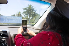A person inside a car is taking a photo or video with a smartphone towards the outside. The individual is wearing a graduation cap and a red outfit with floral patterns. Outside, palm trees are visible, suggesting a sunny location. A banner with the word 'GRADUATES' is visible through the windshield.