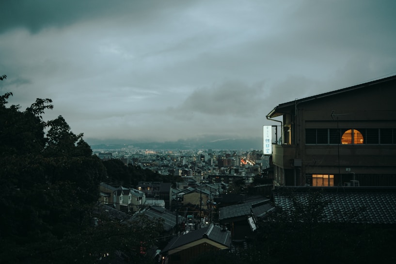 A moody cityscape during twilight, featuring a hillside view overlooking a densely packed urban area. The sky is overcast with heavy, brooding clouds. A prominent building is visible on the right, with warm light glowing from a circular window, creating a stark contrast against the dim surroundings.