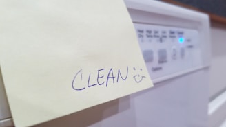 A handwritten note on a yellow sticky paper with the word 'CLEAN' followed by a smiley face. The note is stuck to the front panel of a household appliance, likely a dishwasher, with visible control buttons and a glowing blue indicator light.