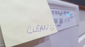 A handwritten note on a yellow sticky paper with the word 'CLEAN' followed by a smiley face. The note is stuck to the front panel of a household appliance, likely a dishwasher, with visible control buttons and a glowing blue indicator light.