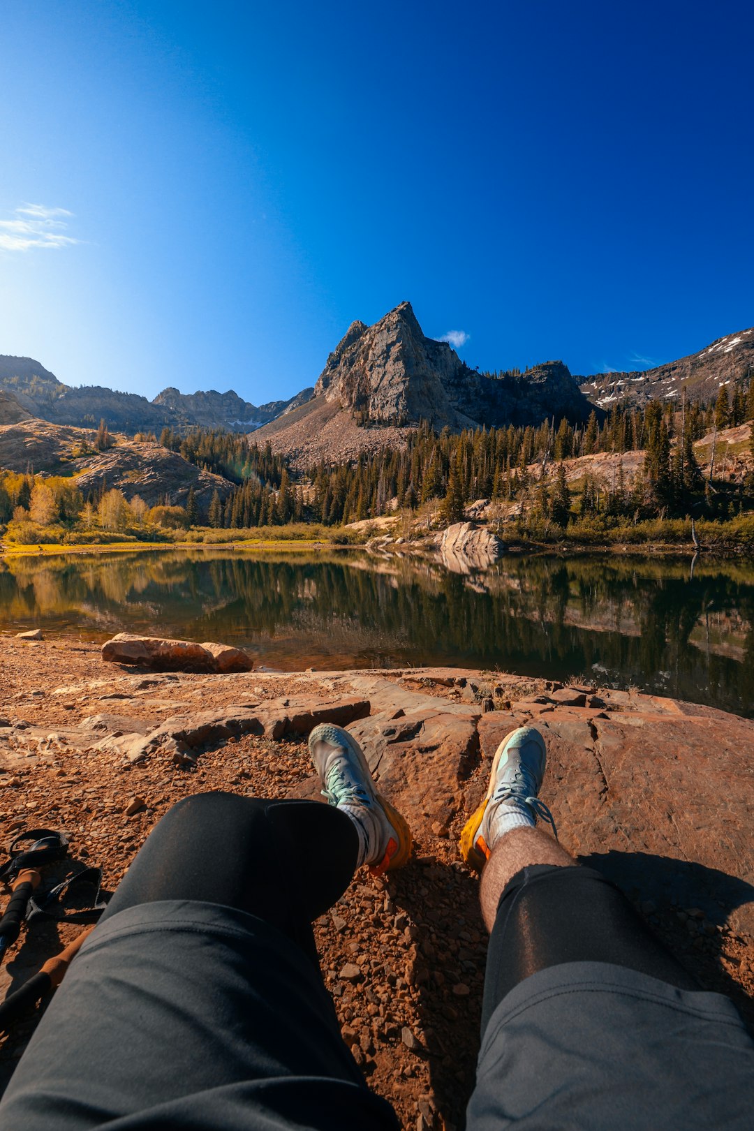 person in black pants and gray sneakers sitting on rock near lake during daytime