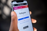 A person is holding a smartphone displaying the Instagram login screen. The phone screen shows the Instagram logo and options to log in or sign up using an email or phone number. The background is blurred and indistinct, focusing attention on the phone.