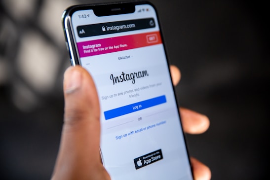 A person is holding a smartphone displaying the Instagram login screen. The phone screen shows the Instagram logo and options to log in or sign up using an email or phone number. The background is blurred and indistinct, focusing attention on the phone.