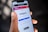 A person is holding a smartphone displaying the Instagram login screen. The phone screen shows the Instagram logo and options to log in or sign up using an email or phone number. The background is blurred and indistinct, focusing attention on the phone.