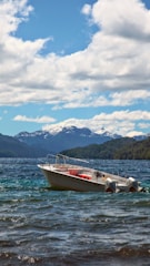 A motorboat is floating on a picturesque lake with clear waters. Snow-capped mountains provide a striking backdrop, and a sky filled with fluffy white clouds adds to the serene atmosphere.