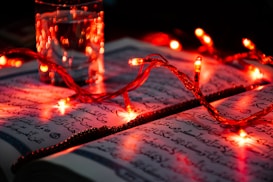 An open book with Arabic script is illuminated by red string lights. A glass of water is placed on the open pages, casting reflections and enhancing the warm glow from the lights.