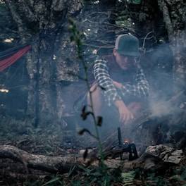 A person wearing a baseball cap and flannel shirt is crouching by a small campfire in a dense forest. Smoke from the fire creates a hazy atmosphere. Nearby, a hammock is tied between trees. The setting is rustic with natural elements such as logs, branches, and foliage surrounding the scene.