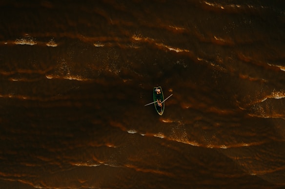 A single canoe is floating on dark water, with visible ripples and textures suggesting movement. The canoe holds a person who appears to be rowing, providing a dynamic element amidst the stillness of the surrounding water.