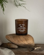 A brown glass candle labeled 'Dry Hay No. 1 Scented Candle' is placed on two large smooth stones. The setup is on a rustic wooden surface with some burnt matchsticks nearby. Delicate green leaves hang in the background, contributing to the natural and earthy aesthetic.