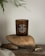 A brown glass candle labeled 'Dry Hay No. 1 Scented Candle' is placed on two large smooth stones. The setup is on a rustic wooden surface with some burnt matchsticks nearby. Delicate green leaves hang in the background, contributing to the natural and earthy aesthetic.