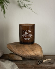 A brown glass candle labeled 'Dry Hay No. 1 Scented Candle' is placed on two large smooth stones. The setup is on a rustic wooden surface with some burnt matchsticks nearby. Delicate green leaves hang in the background, contributing to the natural and earthy aesthetic.