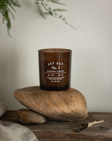 A brown glass candle labeled 'Dry Hay No. 1 Scented Candle' is placed on two large smooth stones. The setup is on a rustic wooden surface with some burnt matchsticks nearby. Delicate green leaves hang in the background, contributing to the natural and earthy aesthetic.