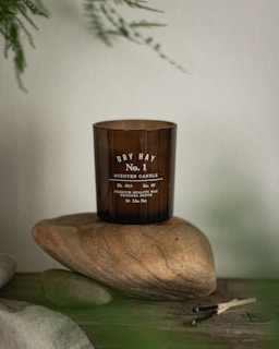 A brown glass candle labeled 'Dry Hay No. 1 Scented Candle' is resting on a smooth, large stone. The surface is wooden and rustic, and there are some burnt matches nearby. In the background, a sprig of greenery is visible.