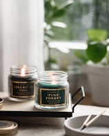 Two lit scented candles in glass jars are placed on a wooden tray. The labels read 'Pine Forest' and 'Firewood Figs'. In the foreground is a speckled ceramic bowl with spent matchsticks. The background shows blurred green potted plants and soft natural lighting, creating a cozy atmosphere.