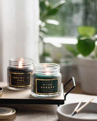 Two lit scented candles in glass jars are placed on a wooden tray. The labels read 'Pine Forest' and 'Firewood Figs'. In the foreground is a speckled ceramic bowl with spent matchsticks. The background shows blurred green potted plants and soft natural lighting, creating a cozy atmosphere.