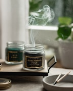 A serene image featuring two glass jars with scented candles on a wooden tray. Smoke gently rises from the burning candle labeled 'A scent of Firewood Figs', creating a calming atmosphere. A small, round dish nearby holds spent wooden matches. In the background, blurred greenery from a potted plant adds a hint of nature.