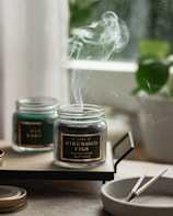 A serene image featuring two glass jars with scented candles on a wooden tray. Smoke gently rises from the burning candle labeled 'A scent of Firewood Figs', creating a calming atmosphere. A small, round dish nearby holds spent wooden matches. In the background, blurred greenery from a potted plant adds a hint of nature.
