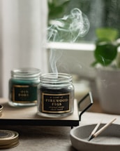 A serene image featuring two glass jars with scented candles on a wooden tray. Smoke gently rises from the burning candle labeled 'A scent of Firewood Figs', creating a calming atmosphere. A small, round dish nearby holds spent wooden matches. In the background, blurred greenery from a potted plant adds a hint of nature.