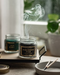 A serene image featuring two glass jars with scented candles on a wooden tray. Smoke gently rises from the burning candle labeled 'A scent of Firewood Figs', creating a calming atmosphere. A small, round dish nearby holds spent wooden matches. In the background, blurred greenery from a potted plant adds a hint of nature.
