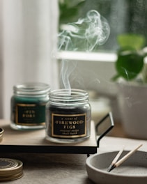 A serene image featuring two glass jars with scented candles on a wooden tray. Smoke gently rises from the burning candle labeled 'A scent of Firewood Figs', creating a calming atmosphere. A small, round dish nearby holds spent wooden matches. In the background, blurred greenery from a potted plant adds a hint of nature.