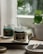 A serene image featuring two glass jars with scented candles on a wooden tray. Smoke gently rises from the burning candle labeled 'A scent of Firewood Figs', creating a calming atmosphere. A small, round dish nearby holds spent wooden matches. In the background, blurred greenery from a potted plant adds a hint of nature.