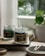 A serene image featuring two glass jars with scented candles on a wooden tray. Smoke gently rises from the burning candle labeled 'A scent of Firewood Figs', creating a calming atmosphere. A small, round dish nearby holds spent wooden matches. In the background, blurred greenery from a potted plant adds a hint of nature.