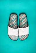 A pair of white slide sandals featuring a textured sole with a mosaic pattern in shades of turquoise, brown, and white, set against a vibrant turquoise background.